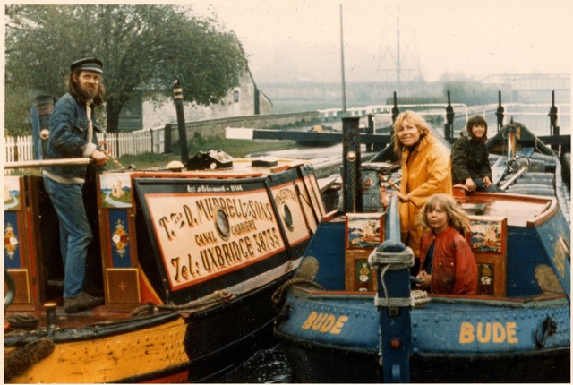 Two canal boats on a river are pictured side by side. The boat on the left bears the slogan "Ted Murrell & Sons" and has a man aboard with a beard and a cap. The boat on the right has two women aboard, one towards the back and one at the front standing across from the man and next to a small child with windswept hair and ruddy cheeks. All are smiling.